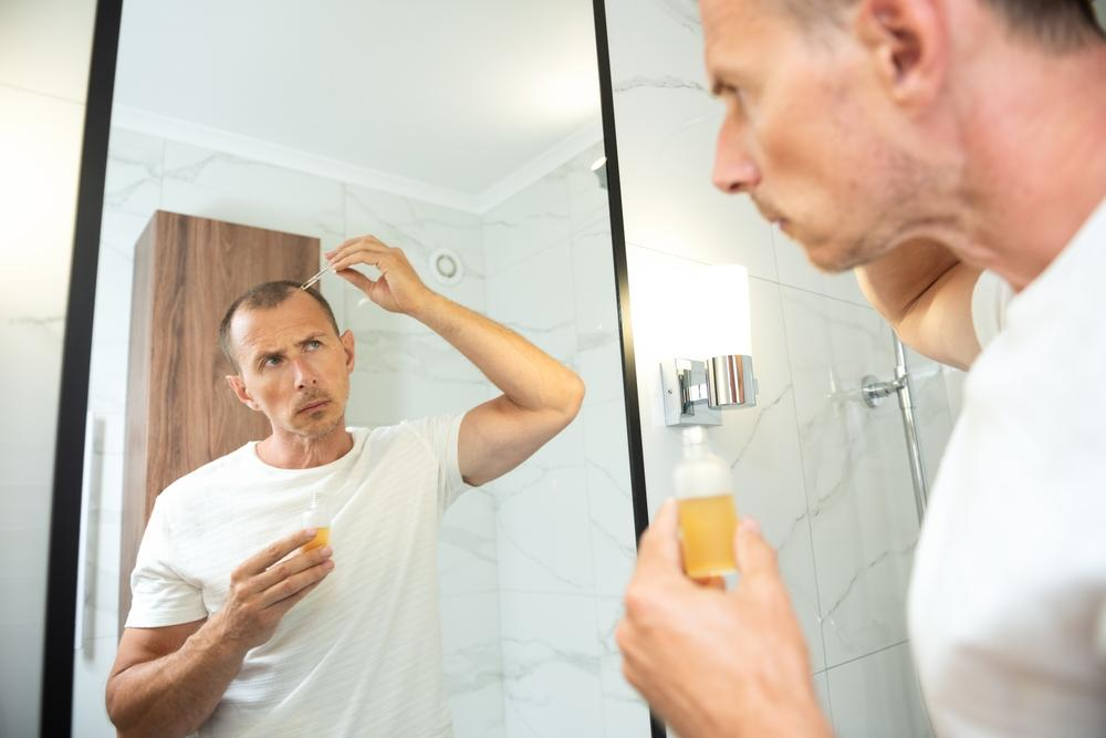Man applying hair treatment to his scalp while looking in the bathroom mirror.